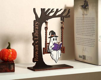 May include: A wooden Halloween decoration depicting a ghost reading a book on a swing. The swing is suspended from a tree with the words "Rowan's Library". A small pumpkin and a book are also visible.