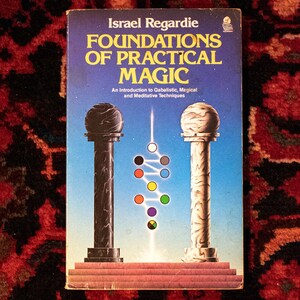 Puede incluir: Un libro azul titulado "Foundations of Practical Magic" de Israel Regardie. La portada del libro presenta una ilustración en blanco y negro de dos pilares con un diagrama de círculos y líneas de colores.