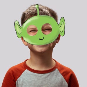 May include: A child wearing a green alien mask with a smiling face and large ears. The mask has eye holes and a small antenna on top.