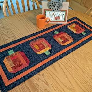 May include: A rectangular fabric table runner featuring three quilted pumpkin motifs in shades of orange, red, and brown, set against a dark blue background with an orange border. An orange ceramic mug and a sign with the text WELCOME sit nearby.