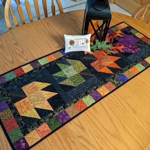 May include: A rectangular quilted table runner featuring a patchwork border of small squares in orange, green, and purple. The center displays large appliqued maple leaves in shades of gold, green, and orange on a black background. A small white rectangular pillow with the text "HELLO autumn" sits on top.