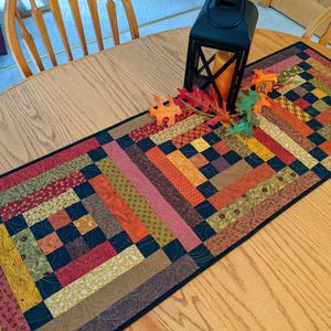 May include: A rectangular quilted table runner with a patchwork design in shades of red, orange, yellow, and green. A black lantern with a candle and decorative fall leaves sit on the runner, which is placed on a wooden table.