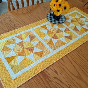May include: A yellow and white quilted table runner with a starburst pattern. The runner has a white border and a yellow patterned edge. A small black and white checkered plate holds a small black cup with yellow flowers.