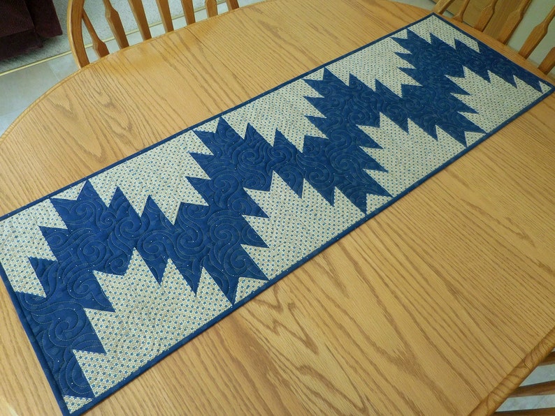 K&ouml;nnte beinhalten: Ein blauer und wei&szlig;er gesteppter Tischl&auml;ufer mit geometrischem Muster. Der L&auml;ufer ist aus Stoff mit einem dezenten Wirbelmuster gefertigt und liegt auf einem Holztisch.