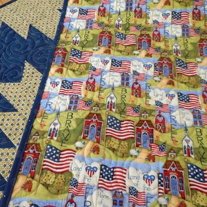 K&ouml;nnte beinhalten: Eine patriotische Steppdecke mit einem blauen und gelben Muster an der Kante. Die Mitte der Steppdecke zeigt ein sich wiederholendes Muster aus rot-wei&szlig;-blauen amerikanischen Flaggen, roten Schulh&auml;usern und gr&uuml;nen B&auml;umen. Der Text "Land of the Free, Home of the Brave" wird auf der gesamten Steppdecke wiederholt.