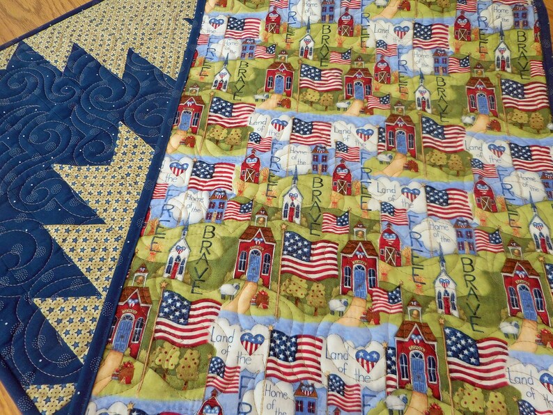 K&ouml;nnte beinhalten: Eine patriotische Steppdecke mit einem blauen und gelben Muster an der Kante. Die Mitte der Steppdecke zeigt ein sich wiederholendes Muster aus rot-wei&szlig;-blauen amerikanischen Flaggen, roten Schulh&auml;usern und gr&uuml;nen B&auml;umen. Der Text "Land of the Free, Home of the Brave" wird auf der gesamten Steppdecke wiederholt.