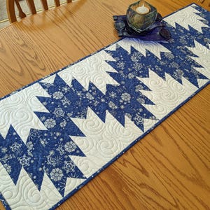May include: A blue and white quilted table runner with a zigzag pattern. The runner features a repeating design of blue snowflake-like shapes on a white background. The runner is approximately 12 inches wide and 48 inches long.