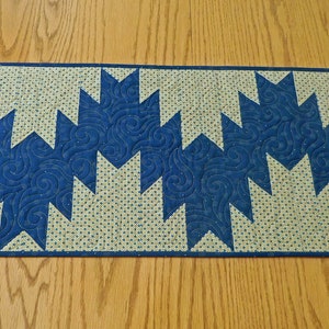 K&ouml;nnte beinhalten: Ein blauer und beiger Tischl&auml;ufer mit geometrischem Muster. Der L&auml;ufer ist aus Stoff und hat eine gesteppte Textur.