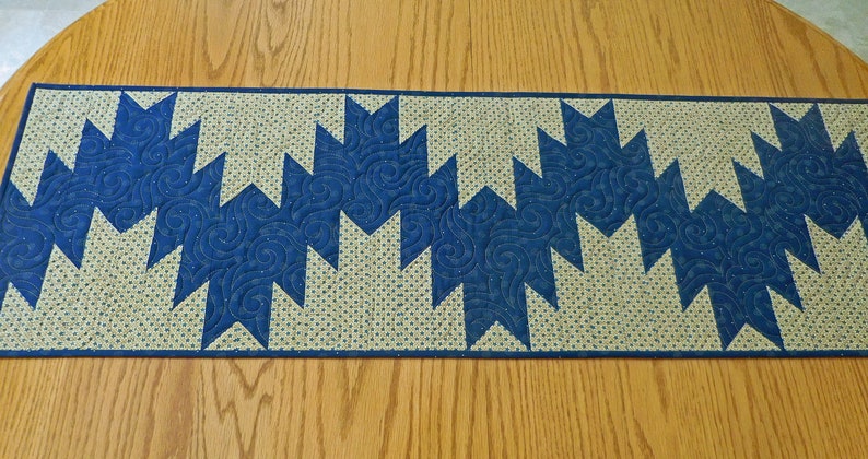 K&ouml;nnte beinhalten: Ein blauer und beiger Tischl&auml;ufer mit geometrischem Muster. Der L&auml;ufer ist aus Stoff und hat eine gesteppte Textur.