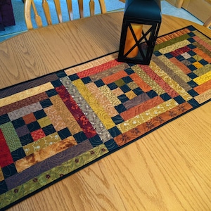 May include: A rectangular quilted table runner featuring a log cabin pattern with various fabric squares and strips in shades of red, orange, yellow, green, brown, and black. The runner has a dark border and intricate machine quilting stitches.