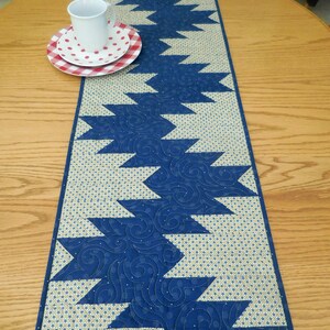 K&ouml;nnte beinhalten: Ein blauer und beiger gesteppter Tischl&auml;ufer mit geometrischem Muster. Der L&auml;ufer hat ein Zickzack-Design mit blauen Dreiecken auf beigefarbenem Hintergrund.