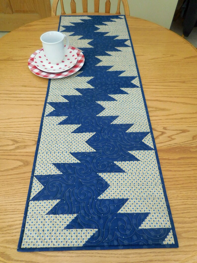 K&ouml;nnte beinhalten: Ein blauer und beiger gesteppter Tischl&auml;ufer mit geometrischem Muster. Der L&auml;ufer hat ein Zickzack-Design mit blauen Dreiecken auf beigefarbenem Hintergrund.