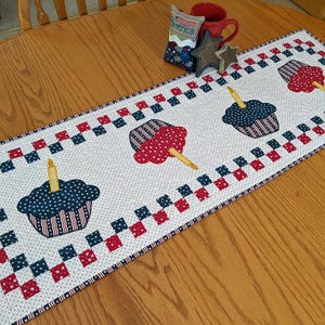 May include: A patriotic table runner featuring cupcake designs with candles. The cupcakes are in red, white, and blue, with a dotted background and a checkered border. A red mug and star-shaped decor are also on the table.