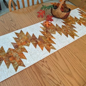 May include: A rectangular quilted table runner featuring a central zigzag pattern of autumn-colored fabric in shades of orange, brown, and gold. The background fabric is cream with a swirling paisley stitch pattern throughout the entire runner.