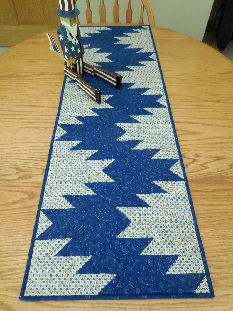 K&ouml;nnte beinhalten: Ein blauer und wei&szlig;er Tischl&auml;ufer mit geometrischem Muster. Der L&auml;ufer ist gesteppt mit einem wirbelnden Design. Eine patriotische Holzfigur sitzt auf dem L&auml;ufer.