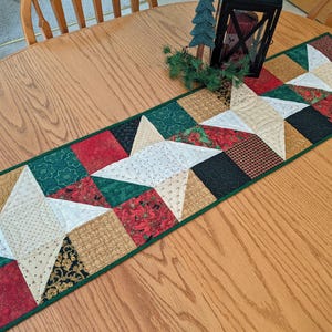 May include: A rectangular quilted table runner featuring a repeating star pattern. The design incorporates various fabric squares in shades of red, dark green, black, and cream, with intricate gold and floral prints, finished with a dark green border.
