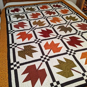May include: A quilt featuring a grid pattern of fabric maple leaves in shades of red, orange, brown, and olive green. The leaves are set against a white background with swirling quilted textures, framed by black and white checkered borders.