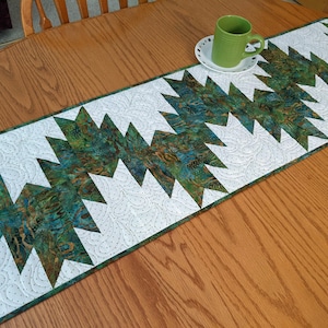 May include: A rectangular quilted table runner featuring a repeating geometric pattern of white fabric with intricate feather stitching and marbled green and blue fabric. A solid green ceramic mug sits on a small white saucer on the runner.