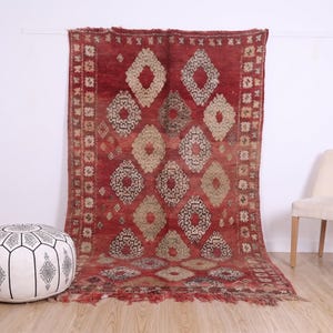 May include: A large rectangular area rug featuring a deep red background with a repeating pattern of beige and dark brown diamond shapes. The rug has a decorative border with small geometric motifs and fringed edges at the top and bottom ends.