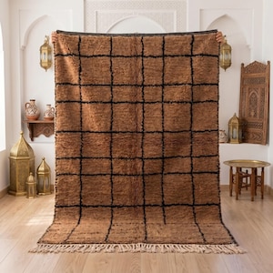 May include: A rectangular area rug featuring a shaggy, textured brown pile with a prominent black grid pattern. The rug has a thick, dark top border and a bottom edge finished with long, light-colored fringe tassels extending from the base.