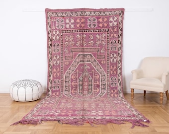 Può includere: Un grande tappeto rettangolare caratterizzato da una base color magenta intenso e viola con intricati motivi geometrici in crema e marrone scuro. Il design include un motivo centrale ad arco, un bordo decorativo e frange annodate.