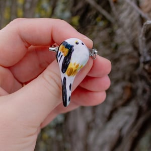 Może przedstawiać: Ręcznie robiona broszka w kształcie ptaka z białą podstawą, zaakcentowana czarnymi, żółtymi i pomarańczowymi detalami. Broszka ma srebrny szpilkę z tyłu. Tło przedstawia rozmyty pień drzewa i gałęzie.