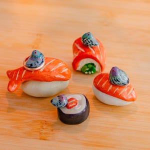 May include: Four miniature ceramic sushi pieces featuring orange salmon and dark seaweed rolls, each topped with a small, hand-painted grey pigeon figurine. The sushi set includes a ring resting on one piece, all displayed on a smooth wooden surface.