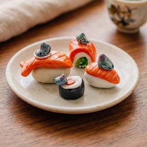 May include: Four ceramic sushi-shaped ring holders on a small, round, off-white plate. Each piece features a miniature gray pigeon figurine perched atop a piece of nigiri or maki sushi, with one silver ring resting on the largest salmon nigiri.