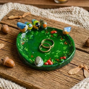 Puede incluir: Un plato decorativo redondo de cerámica verde con un patrón pintado de hojas y bayas en la superficie interior. Seis pájaros pequeños, coloridos y esculpidos están unidos al borde. Dos anillos de metal dorado descansan en el centro.