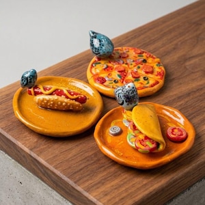Puede incluir: Esculturas de cerámica de comida: un perrito caliente, una pizza y un taco sobre platos naranjas. Cada plato tiene una pequeña escultura azul con forma de pájaro. El perrito caliente tiene mostaza amarilla.