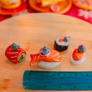 May include: Four ceramic sushi-shaped ring holders, each topped with a small painted bird figurine. The pieces are arranged on a wooden surface next to a blue plastic ruler measuring approximately 12 centimeters in length. The sushi designs include salmon nigiri and maki rolls with vibrant orange, red, and white colors.