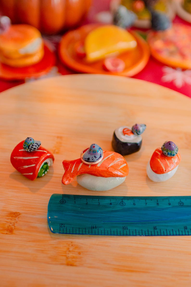 May include: Four ceramic sushi-shaped ring holders, each topped with a small painted bird figurine. The pieces are arranged on a wooden surface next to a blue plastic ruler measuring approximately 12 centimeters in length. The sushi designs include salmon nigiri and maki rolls with vibrant orange, red, and white colors.