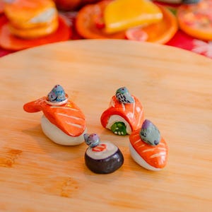 May include: Four ceramic sushi-shaped ring holders, each topped with a small painted pigeon figurine. The set includes two salmon nigiri pieces, one maki roll, and one salmon roll, all featuring orange, white, and black details with grey birds.