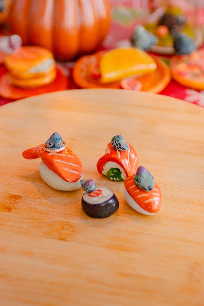 May include: Four ceramic sushi-shaped ring holders, each topped with a small painted pigeon figurine. The set includes two salmon nigiri pieces, one maki roll, and one salmon roll, all featuring orange, white, and black details with grey birds.