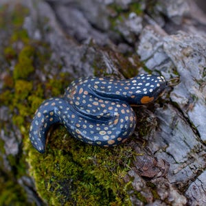 May include: A coiled snake figurine with a glossy dark blue finish and small, light-colored spots across its body. The snake has a smooth, rounded form and a small metal loop attached to its head, suggesting it is designed to be used as a pendant.