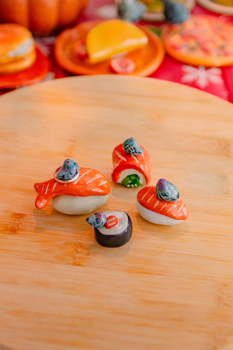 May include: Four miniature ceramic sushi pieces featuring orange salmon and dark seaweed rolls, each topped with a small, hand-painted grey pigeon figurine. The sushi set includes a ring resting on one piece, all displayed on a smooth wooden surface.