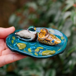 Op de afbeelding: Een handbeschilderde keramische ringschaal met twee eendenbeeldjes op een teal vijver. Eén eend heeft een zilveren ring op zijn rug. De schaal is versierd met gele waterlelies en blauwe waterdetails.