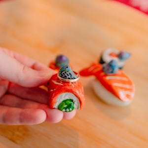 May include: A miniature salmon sushi roll figurine featuring a small gray bird with blue accents perched on top. The sushi roll has a bright orange exterior, white rice, and a green center. A thin metallic ring rests around the bird figure.