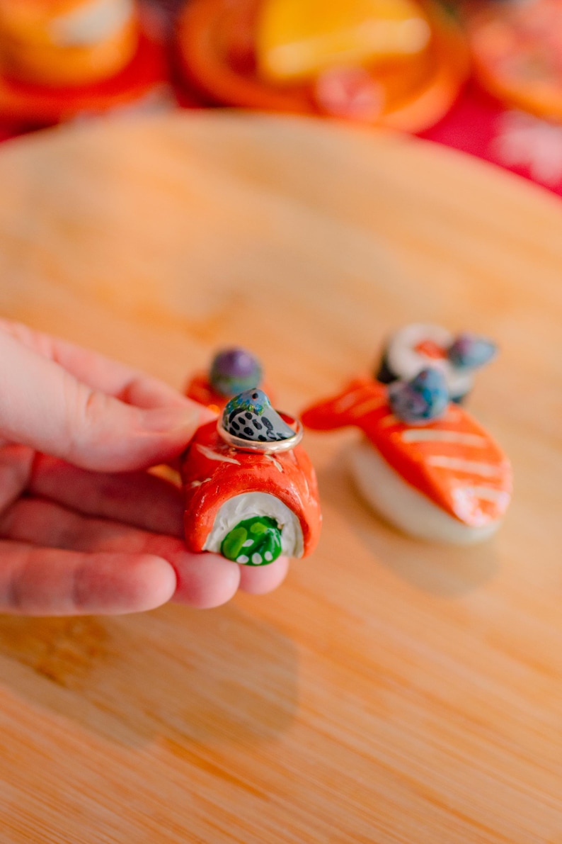May include: A miniature salmon sushi roll figurine featuring a small gray bird with blue accents perched on top. The sushi roll has a bright orange exterior, white rice, and a green center. A thin metallic ring rests around the bird figure.