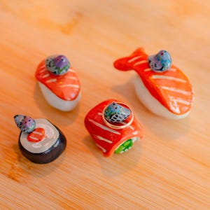 May include: Four ceramic sushi-shaped chopstick rests, each topped with a small, hand-painted blue bird figurine. The set includes two salmon nigiri, one maki roll, and one fish-shaped piece. One nigiri piece features a thin gold ring resting on top.