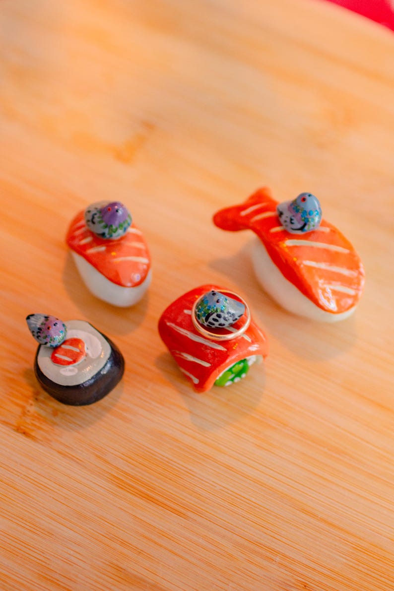 May include: Four ceramic sushi-shaped chopstick rests, each topped with a small, hand-painted blue bird figurine. The set includes two salmon nigiri, one maki roll, and one fish-shaped piece. One nigiri piece features a thin gold ring resting on top.