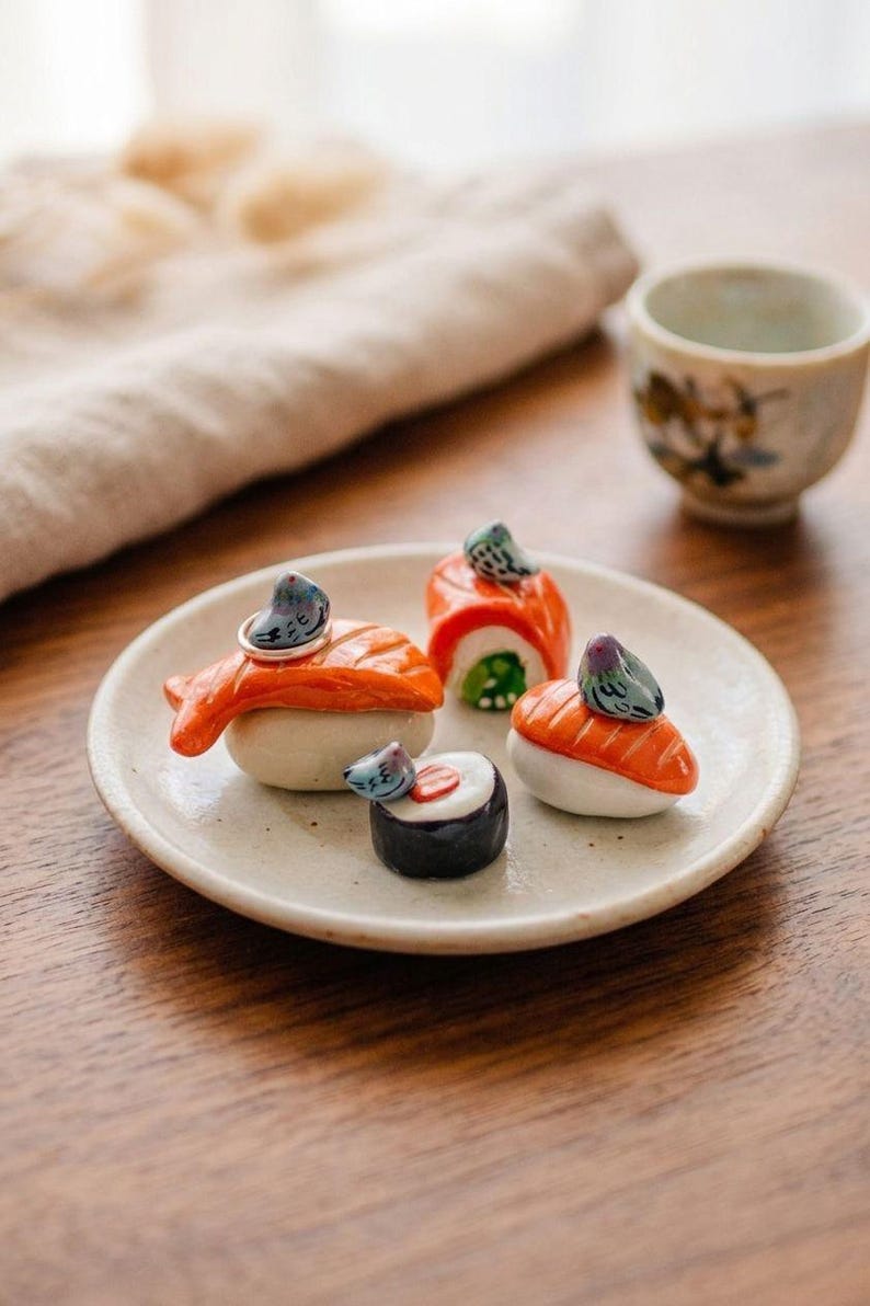 May include: Four ceramic sushi-shaped ring holders on a small, round, off-white plate. Each piece features a miniature gray pigeon figurine perched atop a piece of nigiri or maki sushi, with one silver ring resting on the largest salmon nigiri.