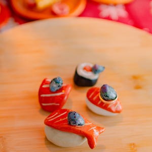 May include: Four ceramic sushi-shaped ring holders featuring orange salmon and white rice textures. Each piece is topped with a small, hand-painted gray bird figurine. The set includes three nigiri-style pieces and one maki roll, designed to hold rings.