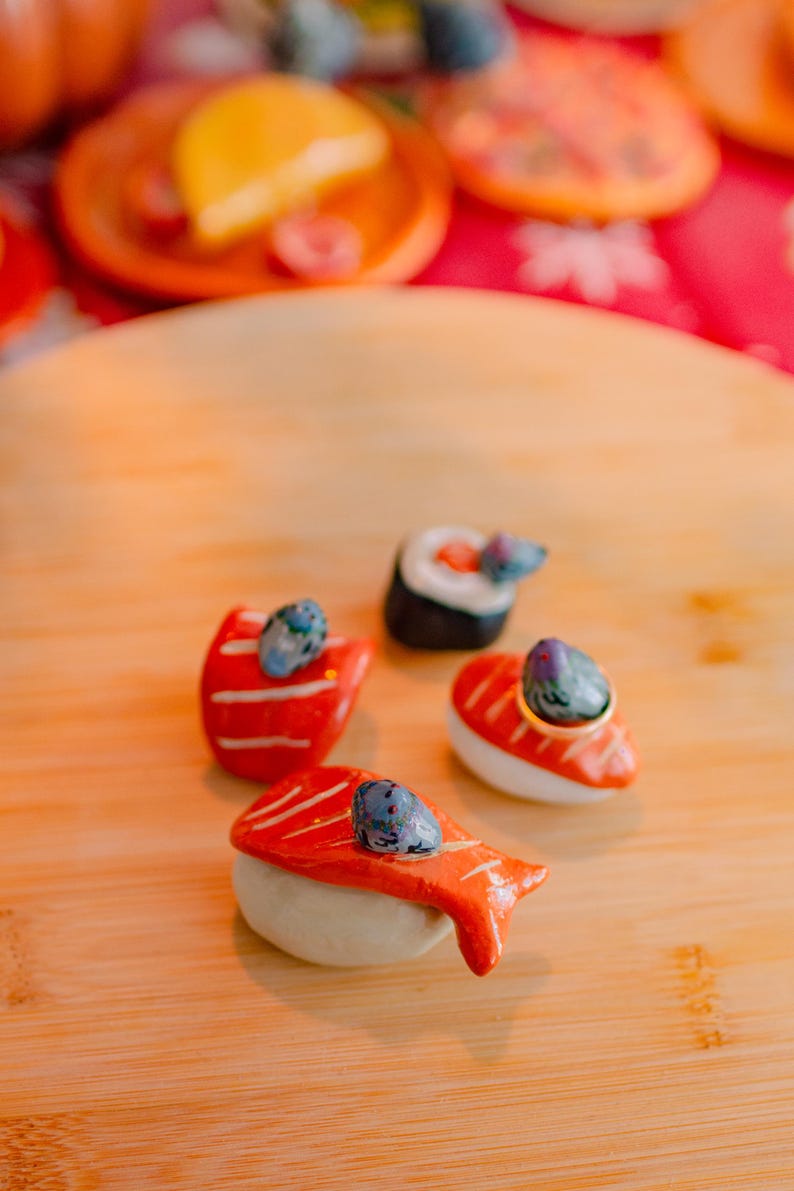 May include: Four ceramic sushi-shaped ring holders featuring orange salmon and white rice textures. Each piece is topped with a small, hand-painted gray bird figurine. The set includes three nigiri-style pieces and one maki roll, designed to hold rings.