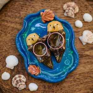 Può includere: Un porta anelli in ceramica blu a forma di due lontre marroni che si tengono per mano, ognuna con un muso beige liscio. Le lontre sono posizionate su una base blu lucida con accenti di glitter verde, progettata per sostenere due anelli.