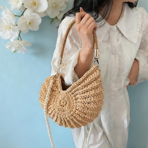 Puede incluir: Un bolso de mano de paja tejida con un asa trenzada y herrajes plateados. El bolso presenta una correa de cadena de perlas y un diseño circular en la parte delantera. El bolso es de color beige y tiene un aspecto texturizado único.