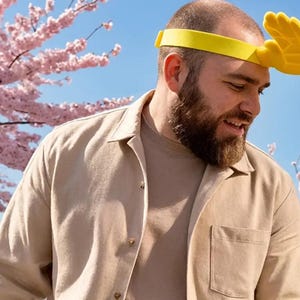 May include: A yellow headband with a hand-shaped design. The person in the image is wearing a tan button-down shirt and a light brown t-shirt. The background features pink flowering trees and a blue sky.
