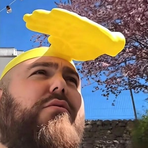 Puede incluir: Un sombrero amarillo brillante con forma de mano, usado en la cabeza. El sombrero es el punto focal, con un cielo azul claro y un árbol floreciente en el fondo. El sombrero es el tema principal.