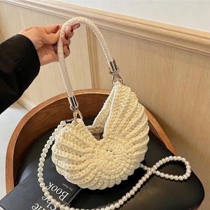 May include: Cream-colored crochet handbag with a braided handle and a pearl-beaded shoulder strap. The bag has a unique, shell-like design with a rounded bottom and a curved top. The handle and metal hardware are silver-toned.