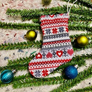 May include: A red and white Christmas stocking with a snowflake and heart pattern. The stocking is surrounded by green pine branches and colorful ornaments.